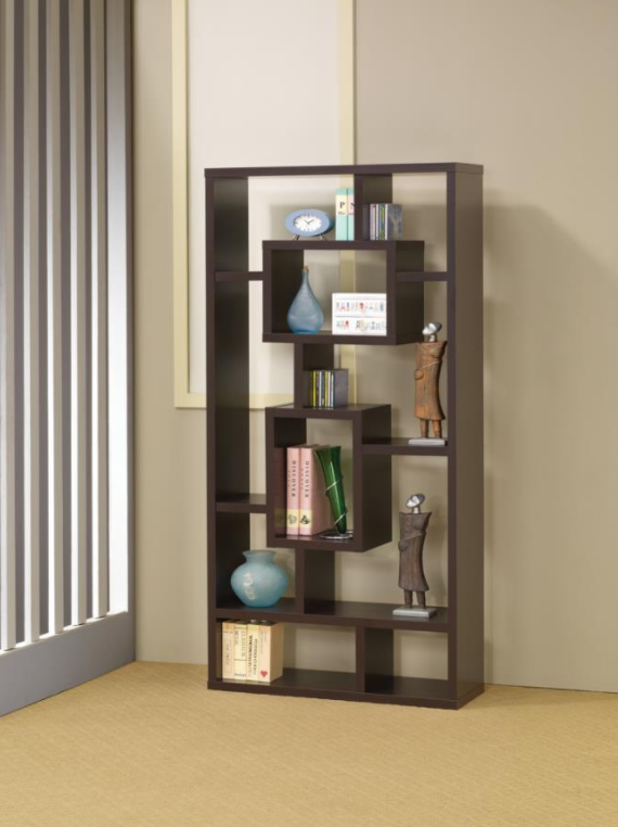 The same brown/cappuccino bookcase in a house setting, with tan walls and carpet. The bookcase features two large rectangular shaped shelves in the center, and the surrounding shelves fill the bookcase in odd tetris-like shapes. The bookcase is riddled with different sized items like large oversized books and a middle sized green vase in 2 of the rectangular shelves. There are also CD case, a small blue alarm clock, and large decorative art pieces that all fit comfortably throughout the bookcase.